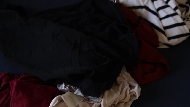 Unfolded clothes lie scattered on a surface as additional garments fall into the frame one by one, thrown by an unseen person. 