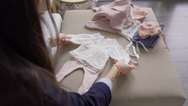 Woman leaning over sofa sorting and arranging knitted baby clothes in pink and white, checking garments before arrival, soft indoor light