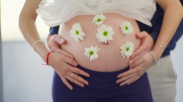 Two pairs of hands holding pregnant belly covered with white daisy flowers, cinematic slider shot, soft indoor light, intimate couple moment