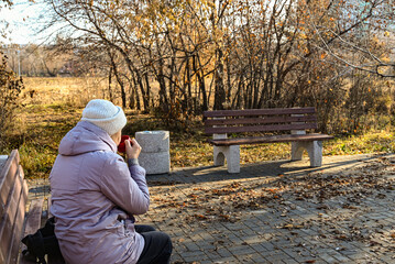 Fototapeta premium Older woman enjoys warm drink on a park bench during autumn afternoon