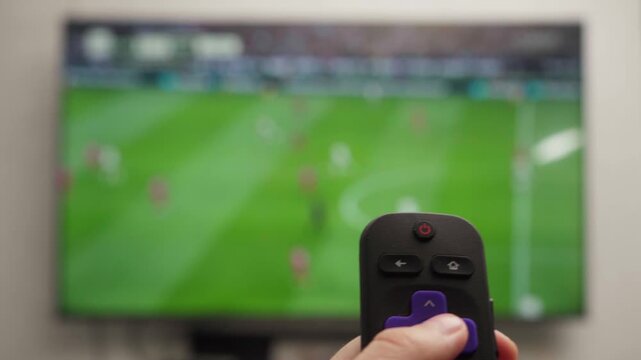 Person watching a live soccer game on smart tv, pointing a remote control at the screen and changing the channel, focusing on the remote with the television out of focus