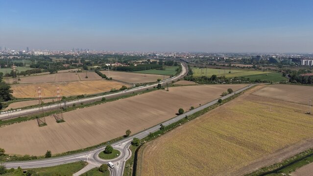 An expansive aerial perspective captures a network of roads gracefully weaving through expansive fields under a clear blue sky, evoking a sense of journey and exploration. San Giuliano Milanese, Lomba