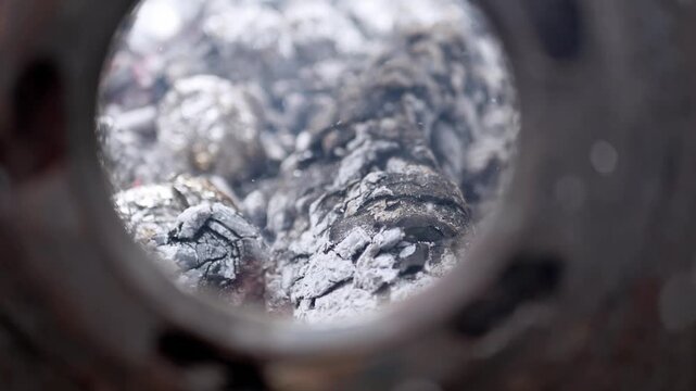 Burning wood logs glow and turn into white ash inside a metal chamber seen through a circular opening. Intense heat and charred textures suggest domestic heating or a dying fire.