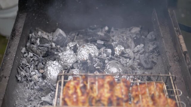 Seasoned chicken meat sears on a metal grill grate while potatoes wrapped in aluminum foil cook slowly within hot glowing charcoal ashes. Concept of rustic weekend outdoor cooking.