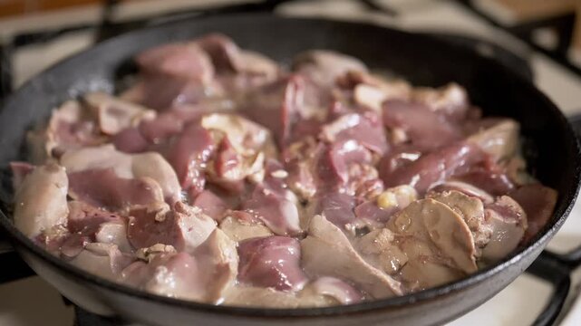 Fresh raw chicken livers are searing in a cast iron skillet on a hot stovetop, browning and sizzling as they cook. Concept of home cooking, traditional offal recipes, and nutrition.