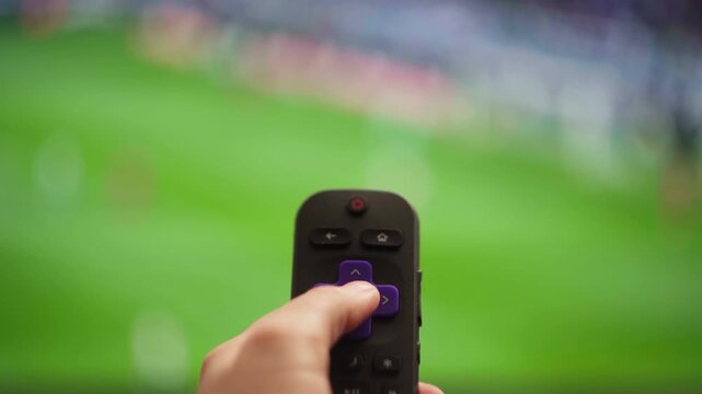 Person using a remote control to change channels on a smart television while watching a soccer match, navigating the on-demand menu and streaming media content
