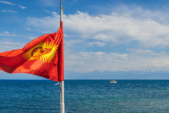 Red flag of Kyrgyzstan waving with boat in serene Issyk-Kul lake water with mountains on the horizon.