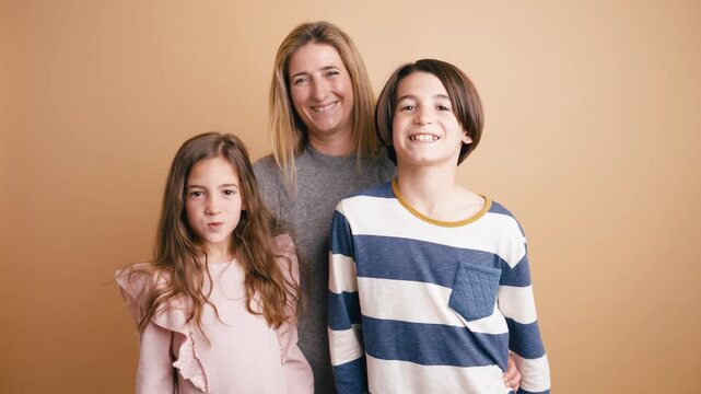 Cheerful mother with her son and daughter are posing together against a plain studio backdrop, smiling and making funny faces