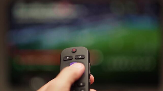 Person changing channels with a smart tv remote control while watching a live sports game, point of view perspective showing the hand holding the electronic device