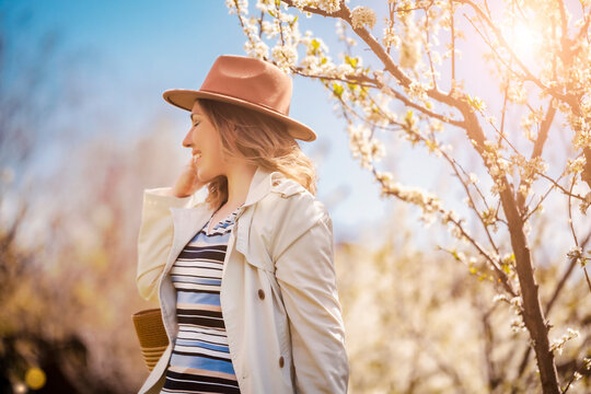 Smiling woman in hat near blooming tree. Peaceful spring portrait and aesthetic appreciation of nature.