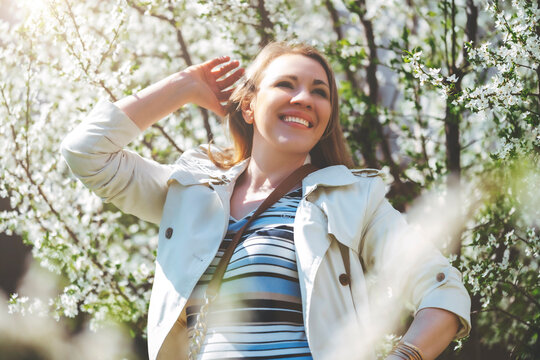Smiling woman touching hair near blooming trees. Graceful spring portrait and internal state of happiness.
