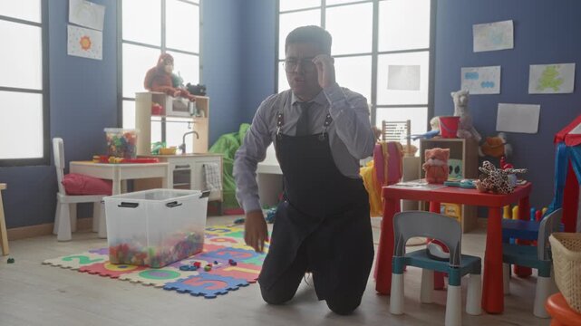 Man teacher kneeling points finger at plastic toy box and alphabet foam mat in kindergarten building giving firm instruction; patience.