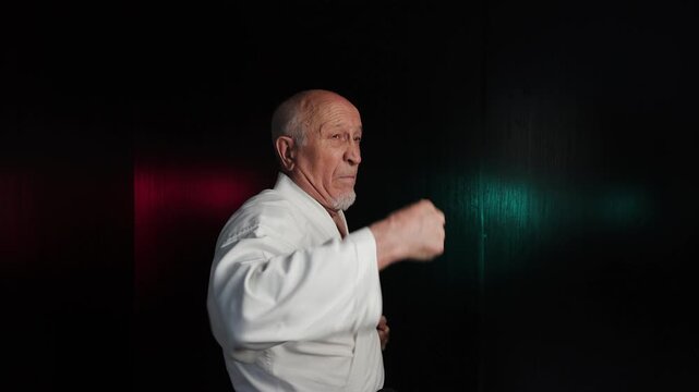 Against a background of flashing red and green lights, an elderly male athlete practices a roundhouse kick with his elbow and fist