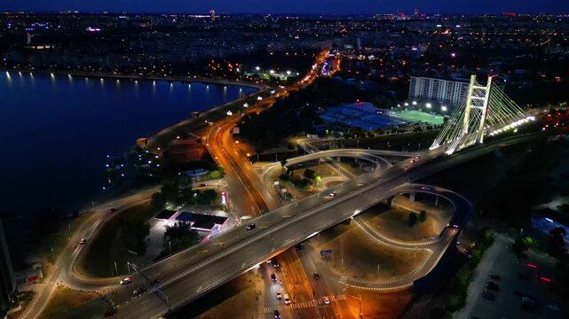 Aerial night drone shot of the illuminated Ciurel bridge in Bucharest, Romania. View of the cable-stayed bridge with traffic light trails and city lights reflecting in the Dambovita river.
