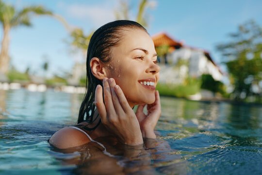 Smiling woman partially submerged in clear pool water cups her face with both hands. Bright outdoor light creates a vibrant, carefree summer mood and a refreshing, relaxed lifestyle scene. promoting