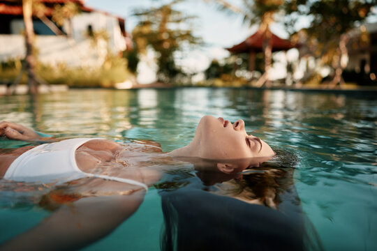 Tranquil woman floats in a clear pool under warm sunlight, enjoying a moment of relaxation. Resort surroundings blur softly in the background, emphasizing wellness and leisure by water.