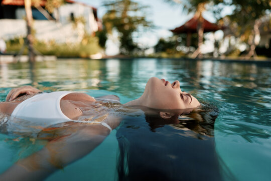 Relaxed person floating on their back in a tranquil pool, sunlit water and a reflective surface, capturing a lifestyle moment of leisure, wellness, and self care, and calm mood.
