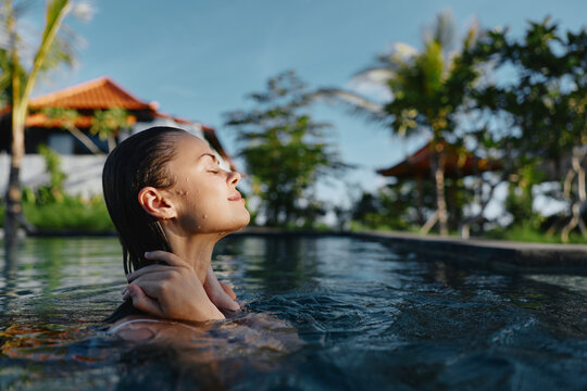 A woman rests in a sunlit pool, eyes closed, savoring relaxation in a tropical outdoor setting, surrounded by palm trees, warm breeze, and clear blue water by a resort.