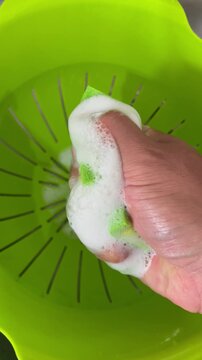 Hand squeezing green sponge with thick white soap foam in basin