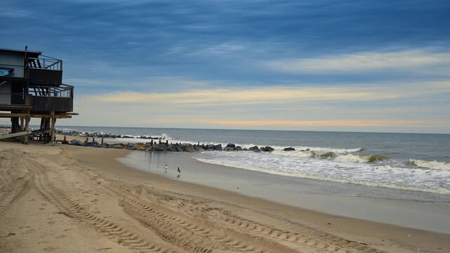 Walk by the sandy beach on a dull cloudy day. Some seagulls fly around. Building on the poles in the backdrop.