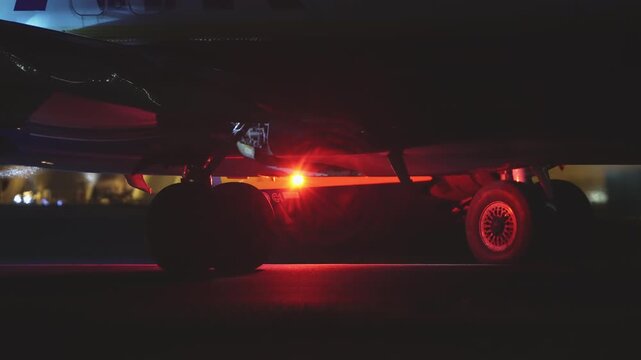 Low angle view of large aircraft underside at night, ground vehicle with yellow green marking visible beneath fuselage, runway light glowing in distance