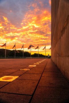 View of the base of the Washington Monument with American flags waving against an orange sunset sky and the distant Capitol building in Washington, District of Columbia, United States.