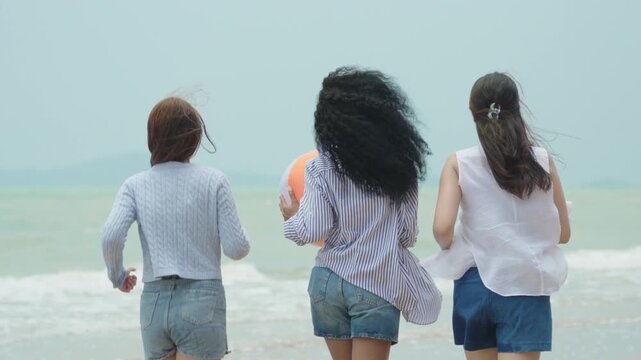 Joyful slow-motion video showing three diverse young women Caucasian, Black, Asian running and jumping together simultaneously on a sandy tropical beach during a sunny summer vacation.
