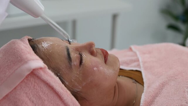 The young woman relaxes during her facial treatment. The expert's hands gently cleanse her eyes, and a soft pink towel caresses her tranquil face. Her relaxed posture conveys comfort and attentive car