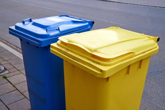 Blue and yellow plastic recycling bins standing on a sidewalk for waste sorting and collection in an urban residential area.