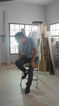 Man sitting on a step ladder with hand on cheek and holding head among packed boxes and bubblewrap in a building during a move; fatigue.