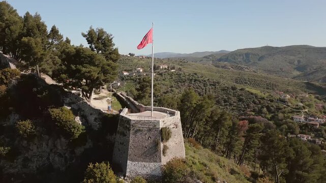 Aerial slow motion view Albanian national flag prominently wave atop stone fortification at Berat Castle - Kalaja e Beratit under clear blue sky, overlooking lush, forested hillside and Berat cityscap