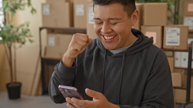Man holding smartphone with fistpump and bright smile amid stacked delivery parcels and shelving in a building, potted plant visible and shipping labels on boxes; small business success.