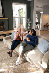 Obraz premium Mature female friends sitting on light sofa in living room, holding ceramic mugs, wearing sneakers