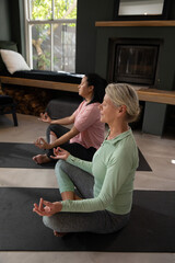 Obraz premium Two mature females in workout clothes sitting cross-legged on black yoga mats at home doing mudra