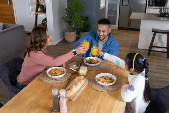 Family with daughter and parents clinking orange juice glasses over pasta bowls at wooden table