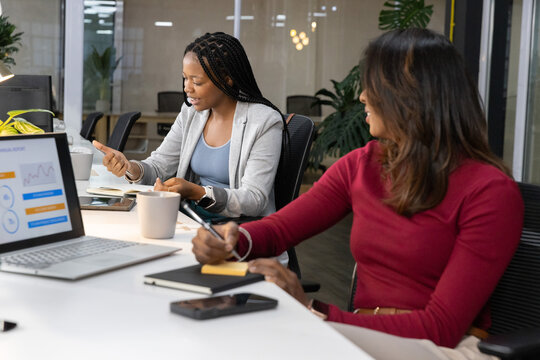 Diverse female coworkers reviewing laptop charts, taking notes at conference table, blazer, red top