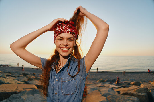 smiling woman with red bandana and denim outfit posing joyfully on rocky beach during golden hour with ocean in background warm sunlight