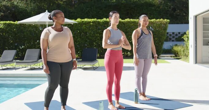 Diverse female friends standing on mats at pool patio, center woman cueing breath into prayer