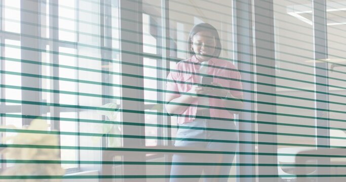 Standing office woman wearing pink jacket and dark skirt holding smartphone at blinds with headset