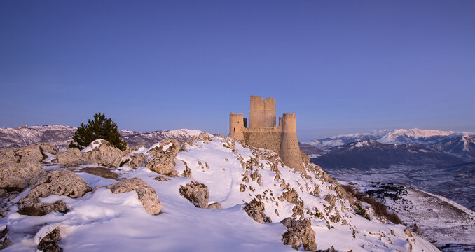 View of the medieval fortress of Rocca Calascio standing on a snow-covered mountain ridge under a clear blue sky in Calascio, Abruzzo, Italy.