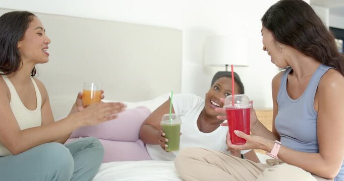 Diverse female friends raising drinks to toast, chatting, clapping, laughing on bed in loungewear