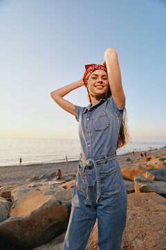 Lifestyle woman in modern boho-western denim street style smiling confidently outdoors near rocky beach with warm film color tone at sunset.