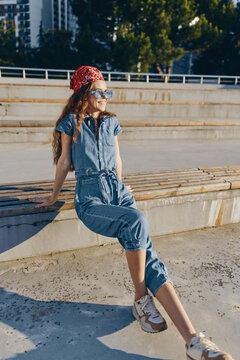 Lifestyle woman wearing modern boho-western denim street style outfit with sunglasses and red bandana, sitting outdoors in warm sunlight, film color effect