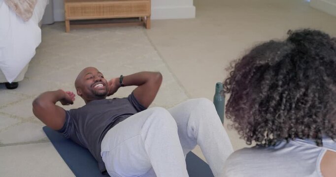 African American man doing sit-ups, woman counting in bedroom on mat, passing can for workout