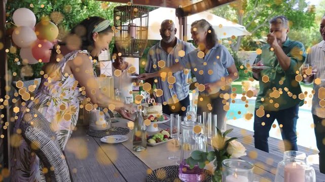 Host carrying cake toward table, guests clapping, gold confetti overlaying scene for birthday party