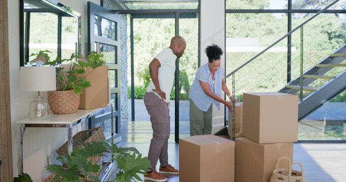 Diverse couple carrying cardboard moving boxes into new home, stacking near staircase and hugging