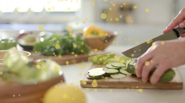 Cooking hands entering, holding, slicing cucumber on wood board, yellow bokeh over hands, prepping