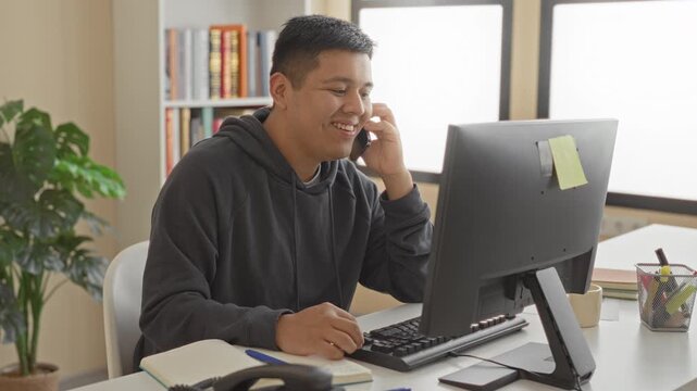 Man holding smartphone to ear and typing on desktop computer at office desk in building with sticky note on monitor and open notebook visible; remote work productivity.