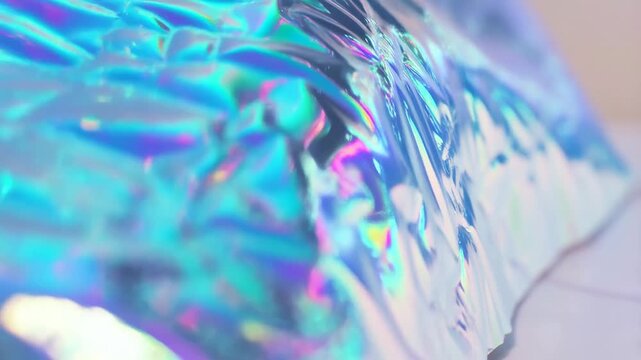 Slow motion tracking shot of iridescent holographic foil crumpling and shimmering on pale tabletop surface in studio closeup