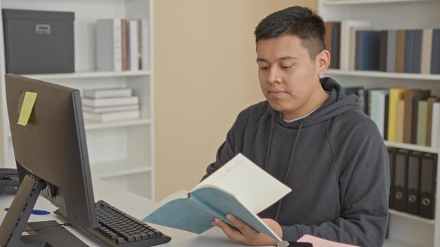 Man reading a blue book, turns page at building study desk with keyboard and monitor, fingers on pages; study concentration.